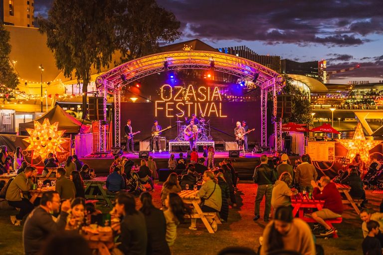 Vibrant night view of the Lucky Dumpling Market at OzAsia Festival in Adelaide, featuring live music on stage and crowds at picnic tables.
